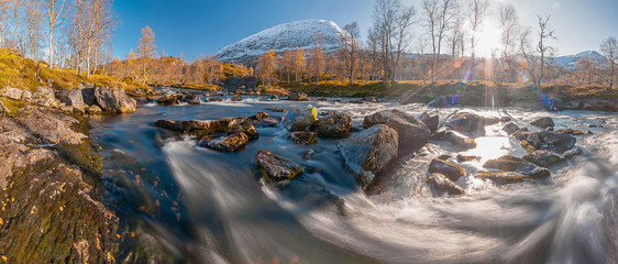 Panoramic view of mountain river flow with motion blurred water. Blue water, golden trees, snow-covered mountains - sunny autumn day in Norway.