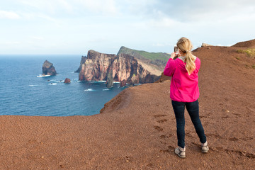 Kobieta blondynka robiąca zdjęcia smartfonem na tle skał i oceanu.