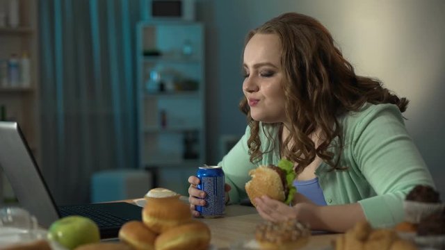 Chubby girl laughing at movie on laptop while eating burger and drinking soda