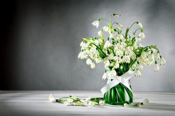 White flowers Loddon Lily stand in a glass vase on white wooden boards. On a gray background.