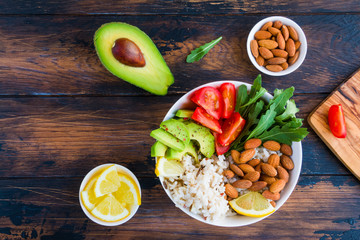 Buddha bowl with avocado, tomato, rice, arugula and almonds. Fresh vegetables, lemon and nuts on the wooden rustic table, top view.