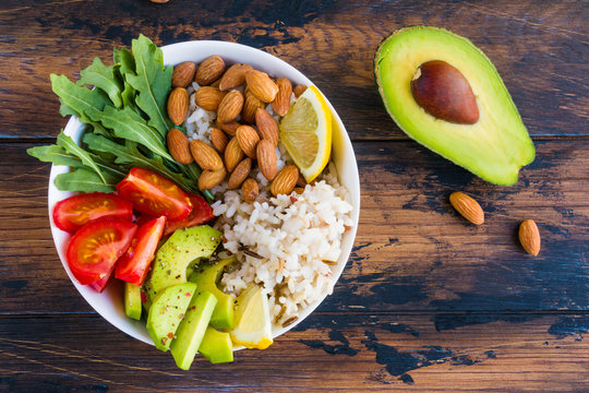 Vegan vegetable Buddha bowl with avocado, tomato, rice, arugula and almonds on the wooden rustic table, top view.