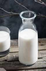 Close-up of a glass of milk and an open glass milk bottle on a rustic wooden table against the dark wall.