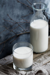 Close-up of a glass of milk and an open glass milk bottle on a rustic wooden table against the dark wall.