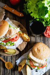 Fast food, barbecue, burger with a white bun with sesame, grilled chicken fillet, fresh cucumber, tomato, lettuce, mayonnaise, with a glass of cola and baked potato on a dark wooden background 