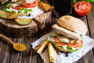 Fast food, barbecue, burger with a white bun with sesame, grilled chicken fillet, fresh cucumber, tomato, lettuce, mayonnaise, with a glass of cola and baked potato on a dark wooden background 