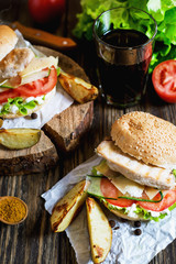 Fast food, barbecue, burger with a white bun with sesame, grilled chicken fillet, fresh cucumber, tomato, lettuce, mayonnaise, with a glass of cola and baked potato on a dark wooden background 