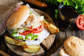 Fast food, barbecue, burger with a white bun with sesame, grilled chicken fillet, fresh cucumber, tomato, lettuce, mayonnaise, with a glass of cola and baked potato on a dark wooden background 