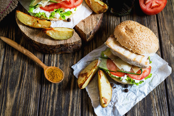 Fast food, barbecue, burger with a white bun with sesame, grilled chicken fillet, fresh cucumber, tomato, lettuce, mayonnaise, with a glass of cola and baked potato on a dark wooden background 