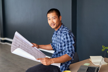 Smiling young Asian architect reading blueprints in an office