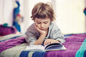 Cute little boy lying on bed reading book