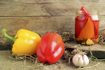 Pickled peppers are in a glass jar. Fresh red and yellow sweet pepper is on a table..