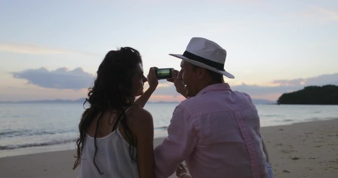 Couple Sitting On Beach Taking Photo Of Sunset On Cell Smart Phone Back Rear View, Young Man And Woman Tourists On Sea Vacation Slow Motion 60