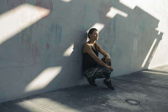 Thoughtful female athlete leaning on wall while crouching at sidewalk