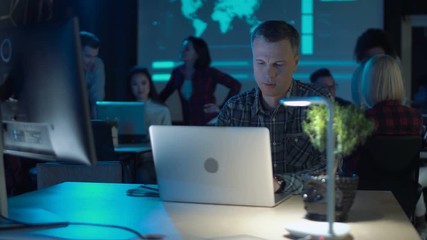 Man at laptop computer working in modern office with low lighting, few of his colleagues are blurred in background. Copy space