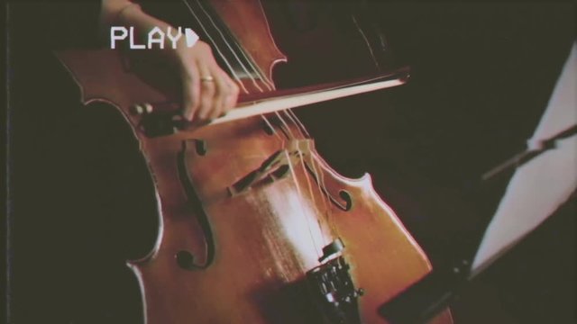 Fake VHS tape: close up of a musician playing a cello in an orchestra.

