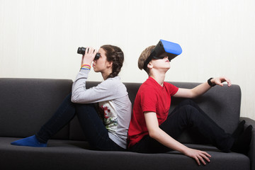 Young girl looking thorough the binocular and the boy wearing virtual reality 3D glasses, sitting on the sofa. Different point of view concept.