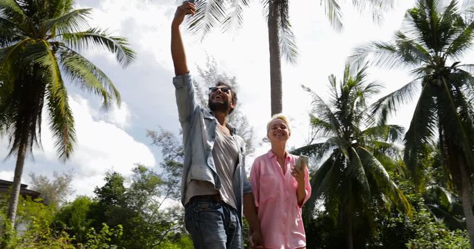 Couple Walk Using Cell Smart Phone Under Palm Trees, Man Hold Hand Up Video Call, Smiling Man And Woman Online Communication Slow Motion 60