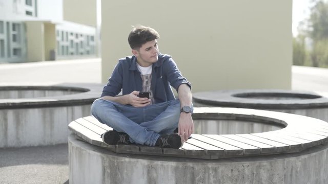 Junger Mann sitzt auf Bank und spielt mit Smartphone