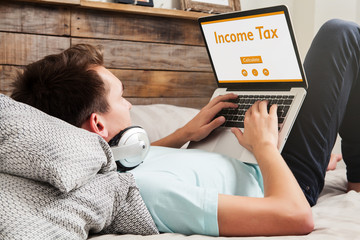 Man doing Income tax declaration on a website with a laptop while lying on the bed at home.