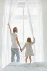 Young mother with daughter opening window curtains in cozy bedroom in the morning. Rear view