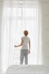 Fototapeta na wymiar Young woman opening window curtains. White color home interior. Rear view