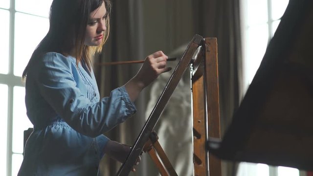 The young artist paints, standing behind an ancient wooden easel. Young woman painter in blue dress paints on canvas in a bright and spacious art workshop. Canvas on the easel.
