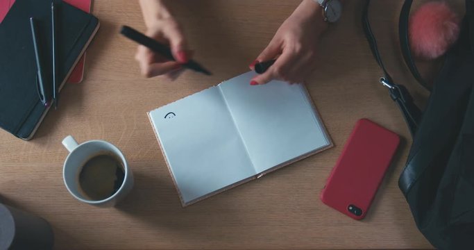 OVERHEAD Female hand drawing a smiling face in opened notebook on wooden table. Top view. Writing concept. Copy space for your text. 4K UHD