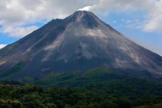 Arenal in Costa Rica. Volcano with exhalation and ash. Beautiful tropic landscape with volcano. Cone active volcano, Central America. Active Arenal with blue sky with clouds. Travelling in Costa Rica