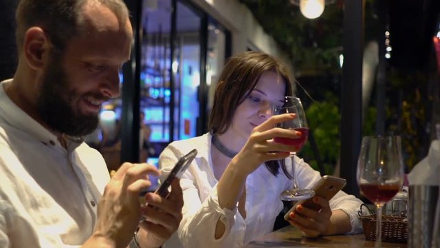 Young couple using smartphones and drinking wine in cafe at night
