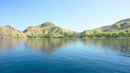 Naklejka na ściany i meble Coastline of mountains with green vegetation reflected in blue ocean water at Labuan Bajo in Flores, Indonesia.