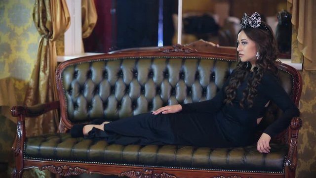Beautiful girl in a sexy long dress, make-up and crown posing for a photographer in a classic interior