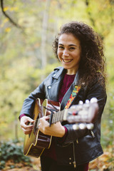 Portrait of beautiful smiling woman playing guitar on forest, fashion lifestyle. Girl wearing black jacket.