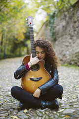 Pensative young woman with a guitar on forest, fashion lifestyle. Girl wearing black jacket.