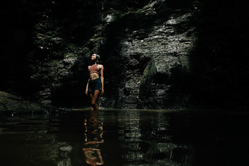Fototapeta premium beautiful wet girl in shorts and a knitted blouse against a waterfall background. Dark background.
