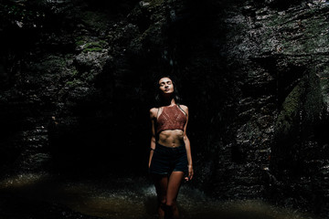 beautiful wet girl in shorts and a knitted blouse against a waterfall background. Dark background.