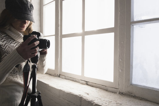 Young woman adjusting camera while standing near window