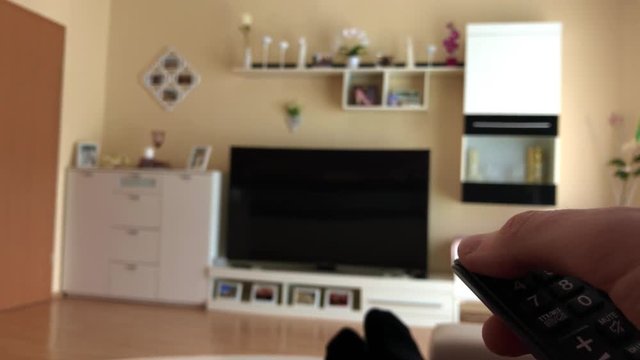 Person sitzt im Wohnzimmer auf Sofa und schaltet Fernseher an bzw. aus