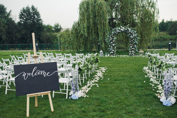 Black board with lettering 'Welcome' stands on the easel on lawn before wedding altar