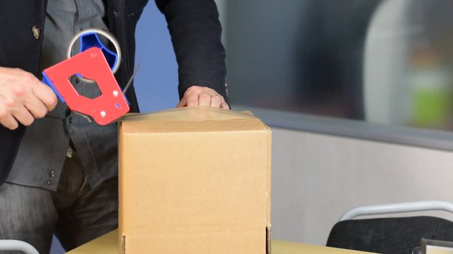 Male hands packing small cardboard box with self-adhesive duct tape in office or warehouse