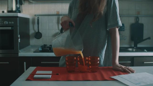 Unrecognizable clumsy brunette woman in grey shirt pouring orange juice out of jug into red glass on kitchen table with notebook and calendar, spills it everywhere