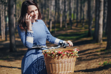 Beautiful girl on a bicycle talking on the phone, with dark hair and hat standing near bicycle with basket .Summer girl enjoying the nature outdoor.Green background.