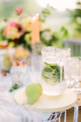 picnic, food, summer, holiday concept - glass jug full of lemonade on festive table, pear, flagon with water, slices of lemon, lime, mint, background of bouquet, candles, glasses
