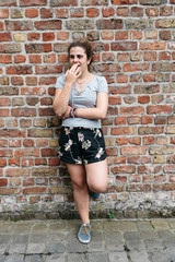 Full length portrait of  young adult woman covering her mouth leaning against brick wall.