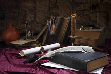 A goose feather, an inkwell, a scroll with a seal, a forged bronze candlestick with a candle, books, a magnifying glass and an hourglass on the desktop. Retro stylized photo. Selective focus