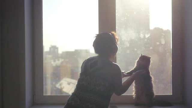 A beautiful cat sits on its hind legs and dances with a girl in headphones against the backdrop of a cityscape with a blur in the sun. slowmotion. 1920x1080