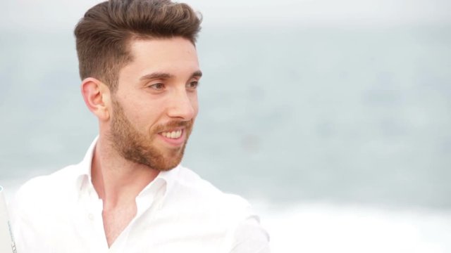 Giovane bello modello con la camicia bianca sorride in riva al mare in Estate. 