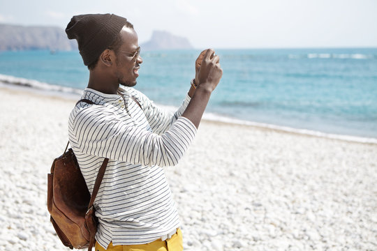 Side view of African American young man with backpack, in hat and striped shirt taking photos of seaside standing on beach alone, posting photo or video on popular social networks during his trip