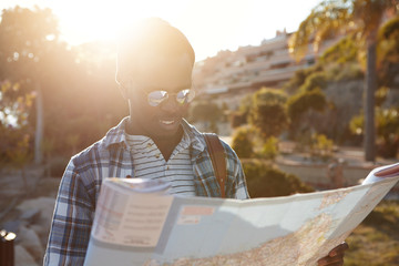 Headshot of cheerful black European guy with backpack traveling alone on foot exploring new beautiful places, holding paper map while looking for old temple in woods. People, travel and adventure