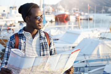 Outdoor portrait of African man looking happy before journey, waiting for his friends in harbor, holding paper map, feeling excited and joyful, anticipating adventures, places and good experience